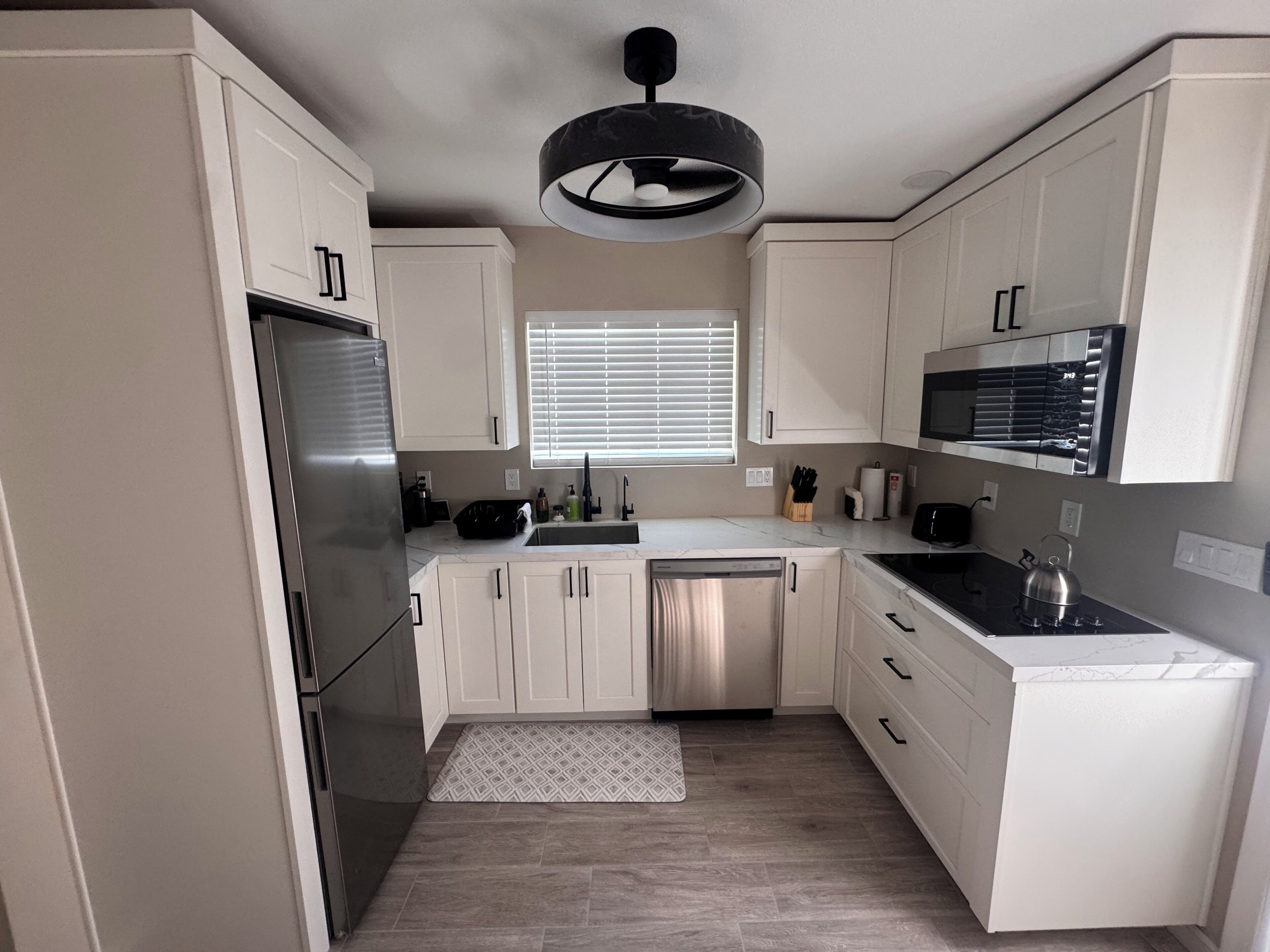 A modern kitchen with white cabinets, stainless steel appliances, a black microwave, and a window with blinds.