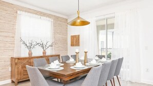 A dining area with a wooden table set for six, grey upholstered chairs, a gold pendant light, and a decorative wall panel.