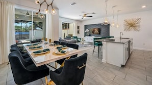 Een moderne eetruimte met een grote tafel voor zes personen, een keuken met een centraal eiland en een zithoek met een bank en een televisie.