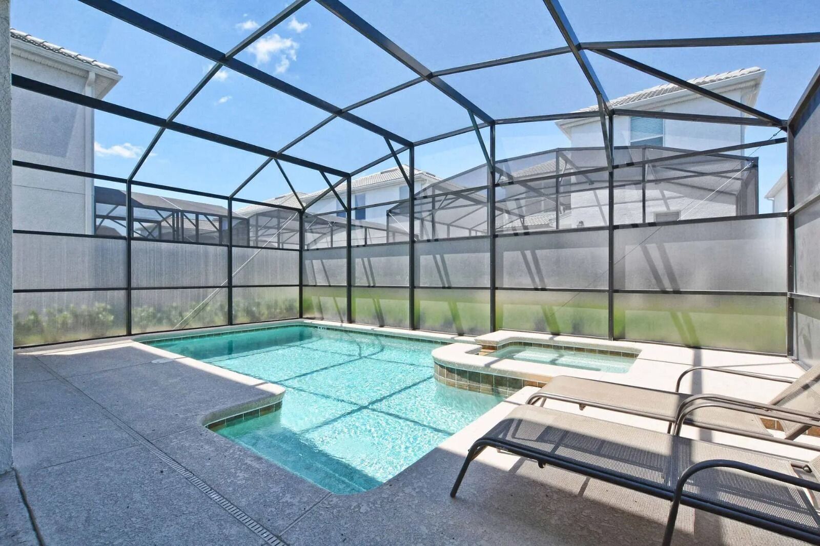 A covered outdoor swimming pool with a hot tub and lounge chairs, enclosed by a metal frame and mesh walls.