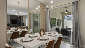 A dining area with a long table set for six, modern chairs, a chandelier, and a view through glass doors to an outdoor seating area. - Themed Home w/ Private Pool (Davenport)