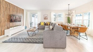 Un salon moderne avec un canapé gris, une table basse et un téléviseur fixé sur un mur en bois. On y trouve également un coin repas avec une table et des chaises en bois, ainsi qu’une grande fenêtre qui laisse entrer la lumière naturelle.