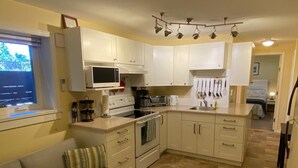 A compact kitchen with white cabinets, a built-in oven, a microwave, and a coffee maker. There is a beige sofa and an ottoman in the living area. - Raven’s Nest – Cozy Lower One‑Bedroom Suite (Victoria)