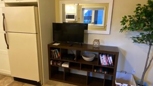 A compact living space with a refrigerator, a television on a wooden stand, a small bookshelf, and a potted plant. - Raven’s Nest – Cozy Lower One‑Bedroom Suite (Victoria)