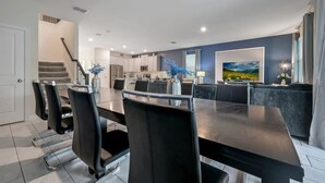 Une cuisine moderne avec des meubles blancs, un coin repas doté d’une grande table et de chaises, et un salon avec un téléviseur et des œuvres d’art accrochées au mur.