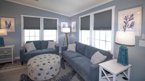 A living room with two grey sofas, a round ottoman, a small white table, and a blue lamp. - Ocean Breeze 2BR/2BA Beach Getaway (Fort Myers Beach)