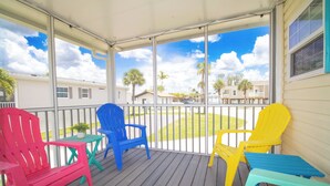 A covered porch with colorful Adirondack chairs and a small table, overlooking a residential area with houses and palm trees. - Ocean Breeze 2BR/2BA Beach Getaway (Fort Myers Beach)