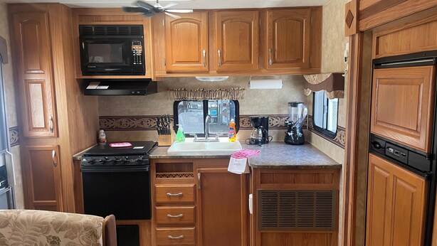 A compact kitchen with wooden cabinets, a microwave, stove, sink, and a ceiling fan.