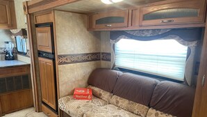 A compact RV interior with a kitchenette, a cushioned seating area, and a window with blinds.