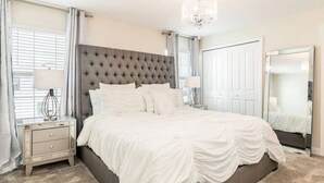 A bedroom with a tufted headboard, a large mirror, a bedside table with a lamp, and a carpeted floor. - Home w/ Private Pool 10 min to Disney (Kissimmee)