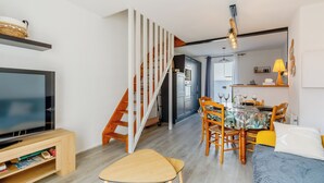 Una sala de estar moderna con un televisor de pantalla plana, escalera de madera, comedor con sillas y mesa, y una cama con colcha azul.