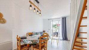 Un comedor con una mesa de madera, sillas y una araña. Se ve una ventana con cortinas y vistas al exterior. A la derecha, se observa una escalera de madera.