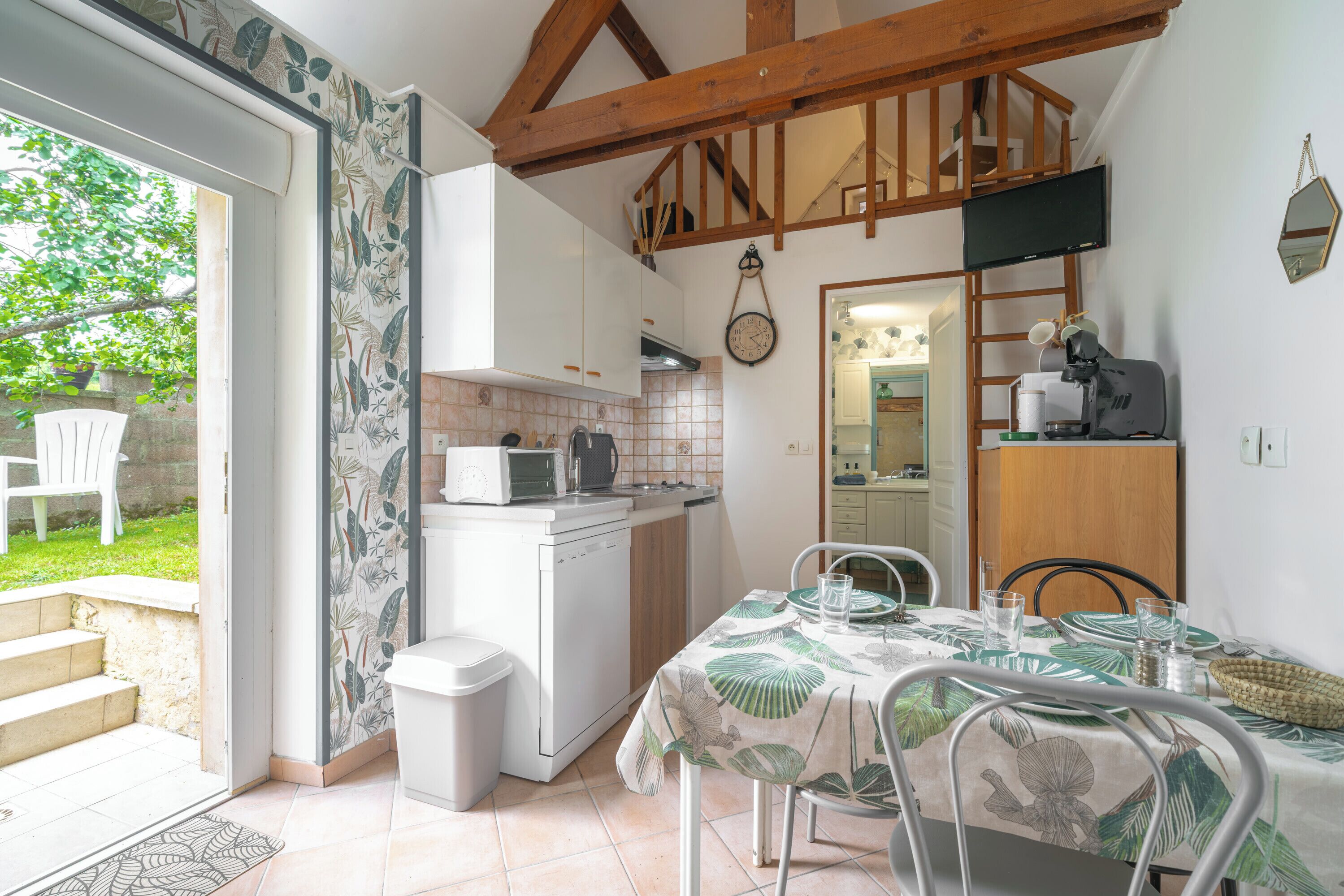 A kitchen with white cabinets, a microwave, and a sink. There is a dining table with chairs and a window overlooking a garden.