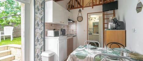 A kitchen with white cabinets, a microwave, and a sink. There is a dining table with chairs and a window overlooking a garden.