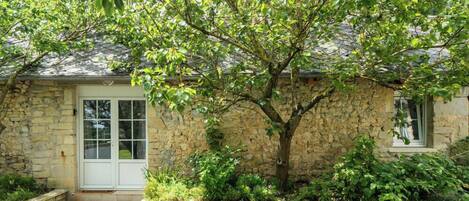 A stone house with a white door, a small porch, and a tree in front.