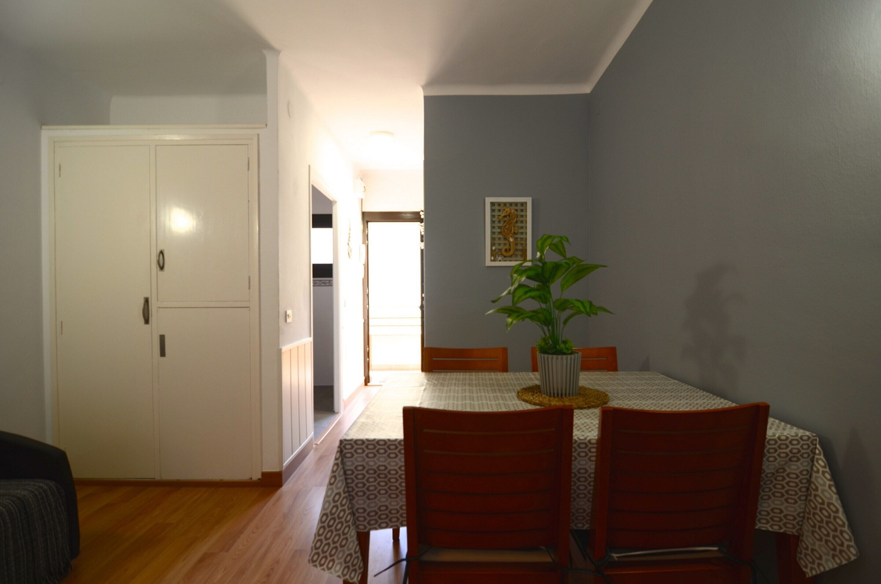 Un comedor con una mesa dispuesta para cuatro personas, una planta en maceta y una puerta blanca.