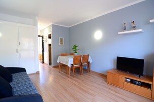 Una sala de estar con un sofá, una mesa de comedor con sillas, un televisor sobre un mueble de madera y un estante con adornos.
