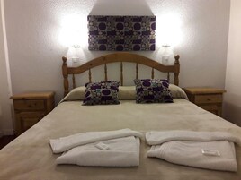 Una cama con cabecera de madera, dos mesitas de noche y dos almohadas con diseño geométrico. Hay dos toallas blancas dobladas sobre la cama.