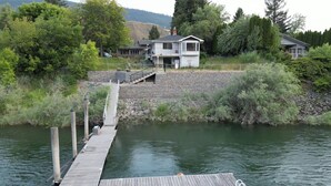 A wooden dock extending into a calm body of water, leading to a house with a stone retaining wall and a covered porch. - Private Room at Riverfront House (Kamloops)