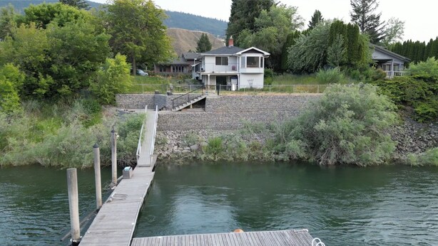 Un moll de fusta s'endinsa en una calma superfície d'aigua, conduint a una casa amb un mur de gabia de pedra i un porxo cobert.