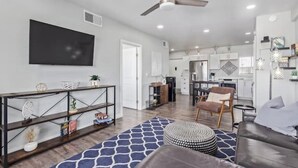 A modern living room with a sofa, ottoman, and a coffee table. There is a TV mounted on the wall and a ceiling fan. - Chic & Stylish Midtown PHX Condo Retreat - Stunning Views & Light Rail Access (Phoenix)