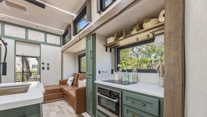 A compact kitchen with a built-in oven, a dining area with a brown leather sofa, and a window with a view of greenery. - Private Lakeside RV Escape with Hot Tub, Multi-Level Deck, & Easy Water Access (Corsicana)