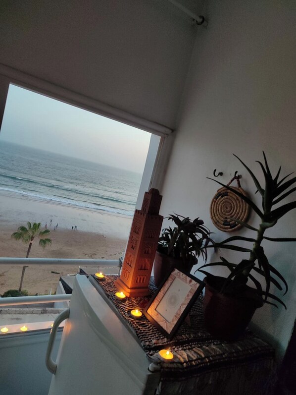 A balcony with a view of the beach, a potted plant, a decorative wall hanging, and a candle holder with lit candles.
