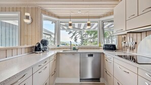Een moderne keuken met houten lambrisering, roestvrijstalen apparatuur en grote ramen met uitzicht op bomen en de zee.