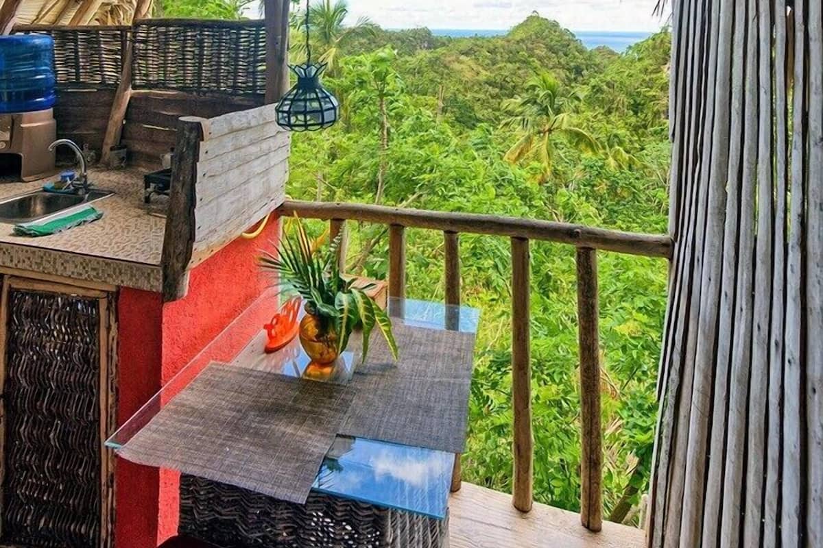 A thatched-roof balcony with a table, chairs, and a view of a lush landscape.