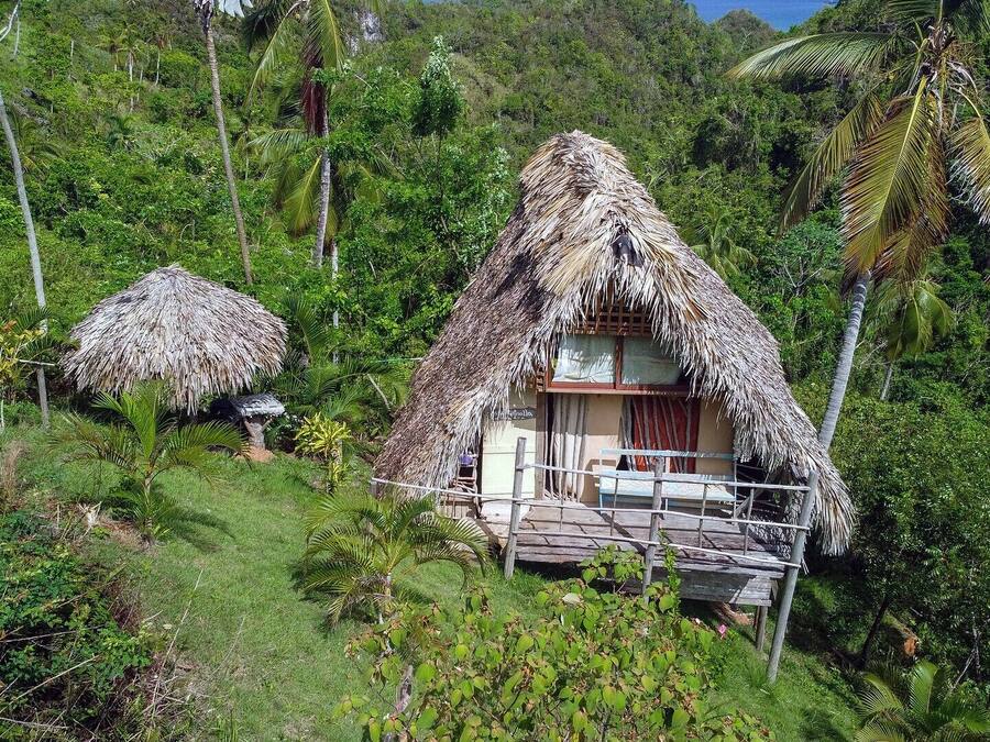 Een rieten huis met een balkon, omringd door weelderige begroeiing en palmbomen.