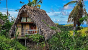 Une cabane à toit de chaume dotée d’un balcon en bois, entourée d’une végétation luxuriante et de palmiers.