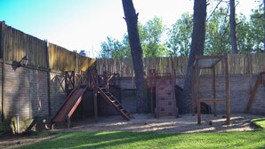 A fenced backyard with a wooden climbing structure, a slide, and a small wooden playhouse. - Alamos Hosteria (Mar Azul)
