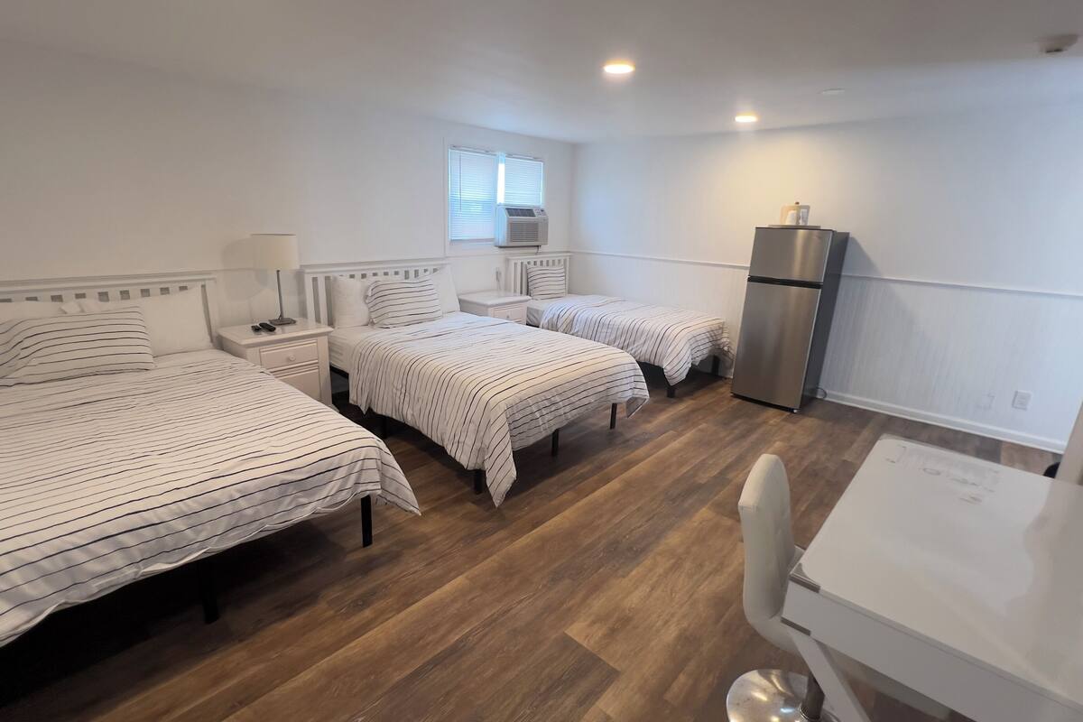 A compact hotel room with two beds, a desk, a chair, a refrigerator, and a window with blinds.