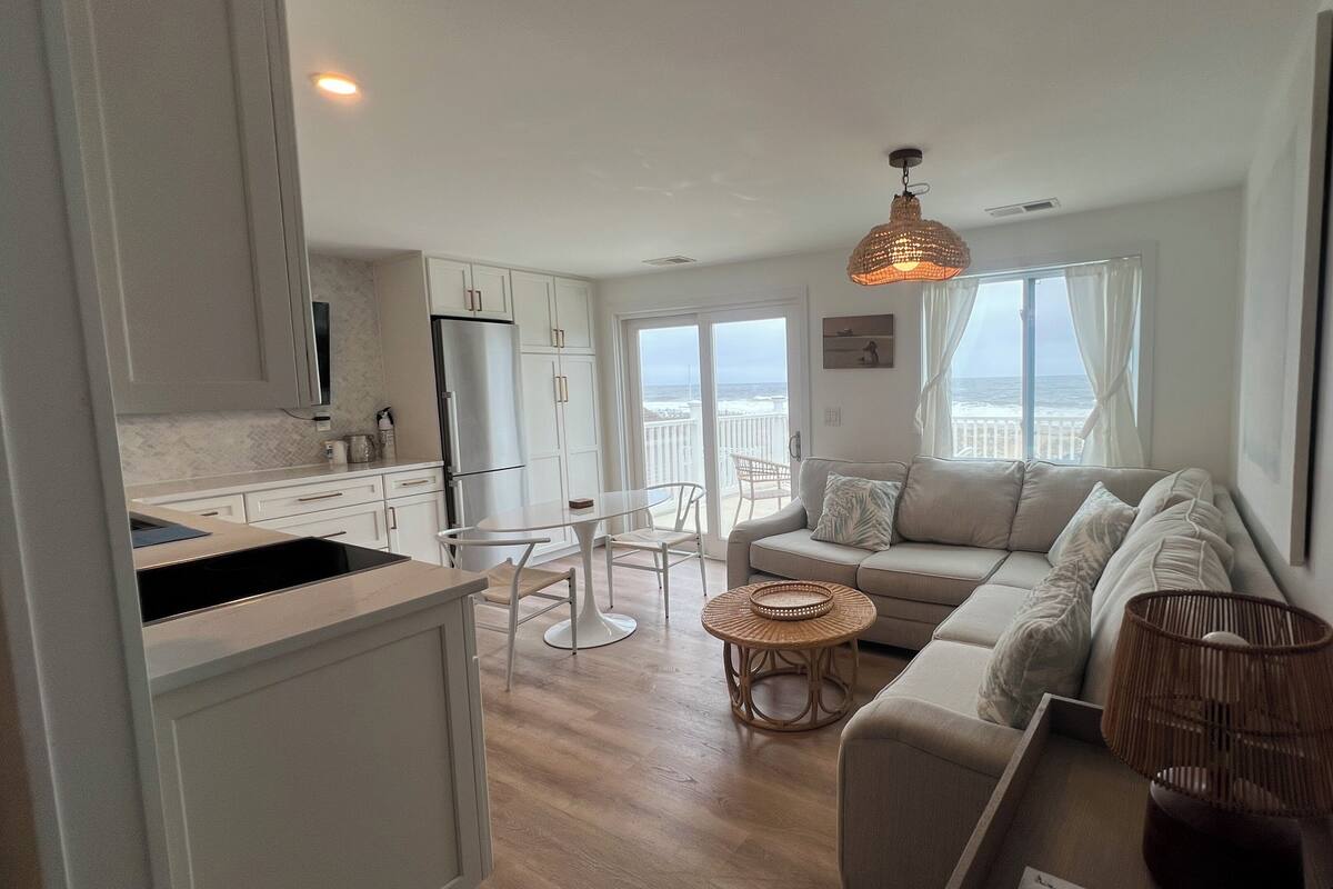 A modern kitchen with white cabinets and a large island, a cozy living area with a sofa and a round coffee table, and a balcony with a view of the sea.