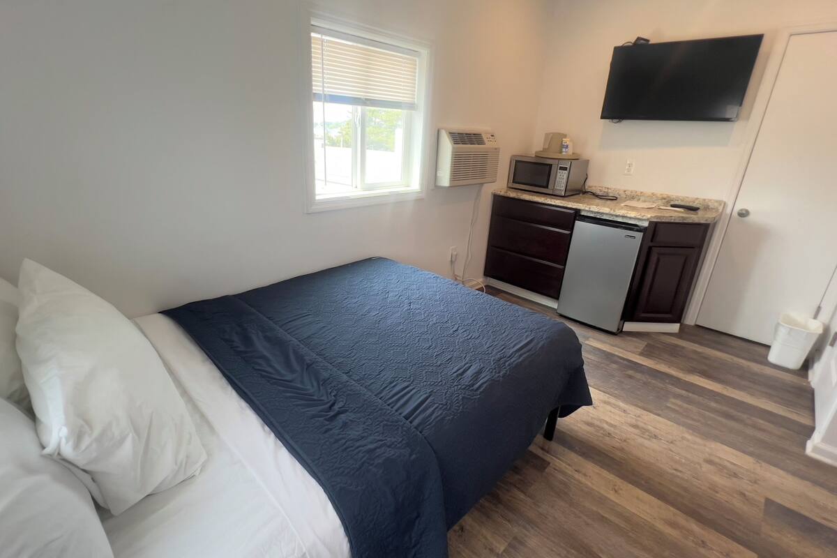 A bedroom with a bed, a small kitchenette with a microwave and refrigerator, and a TV mounted on the wall.
