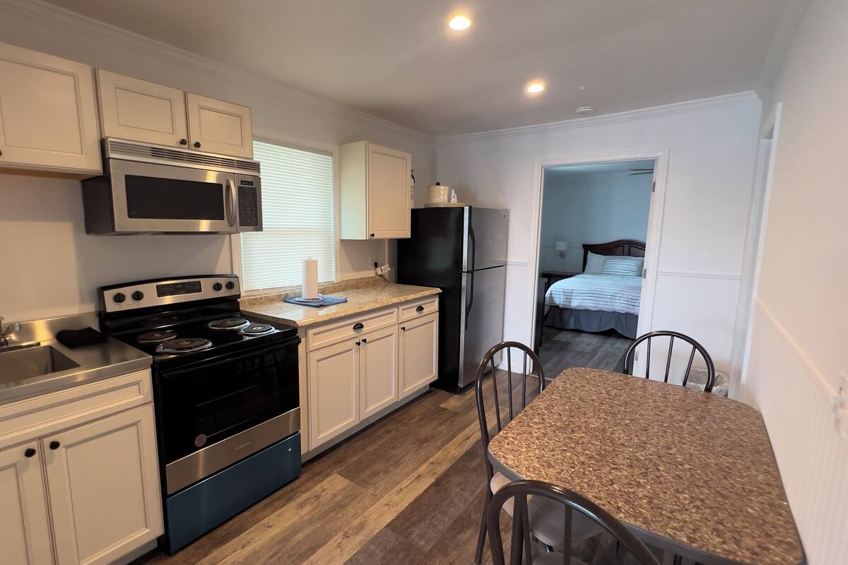 A compact kitchen with white cabinets, a stove, microwave, and refrigerator. There is a dining area with a table and chairs.