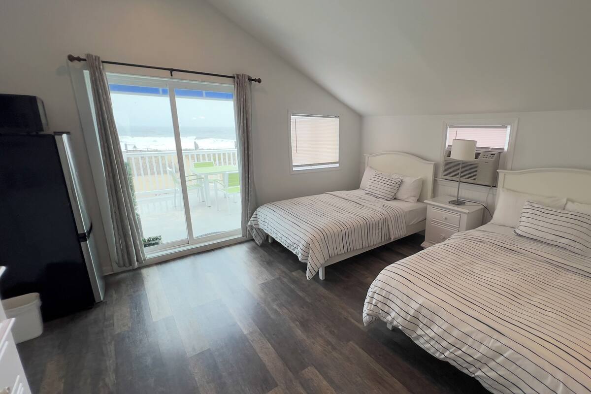 A bedroom with two beds, a nightstand, a window with blinds, and a view of the outdoors.