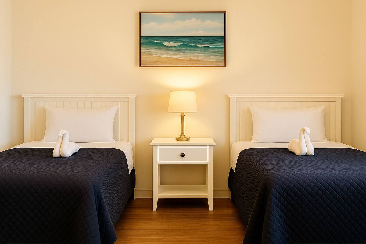 A hotel room with two beds, each with a swan-shaped pillow, a nightstand with a lamp, and a painting of the ocean on the wall.