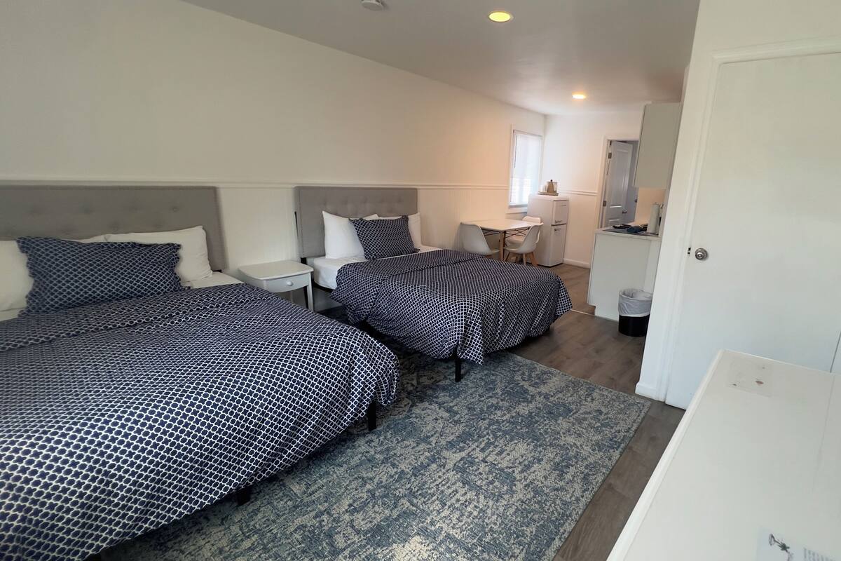 A hotel room with two beds, a desk, a chair, a small table, a mirror, and a door.