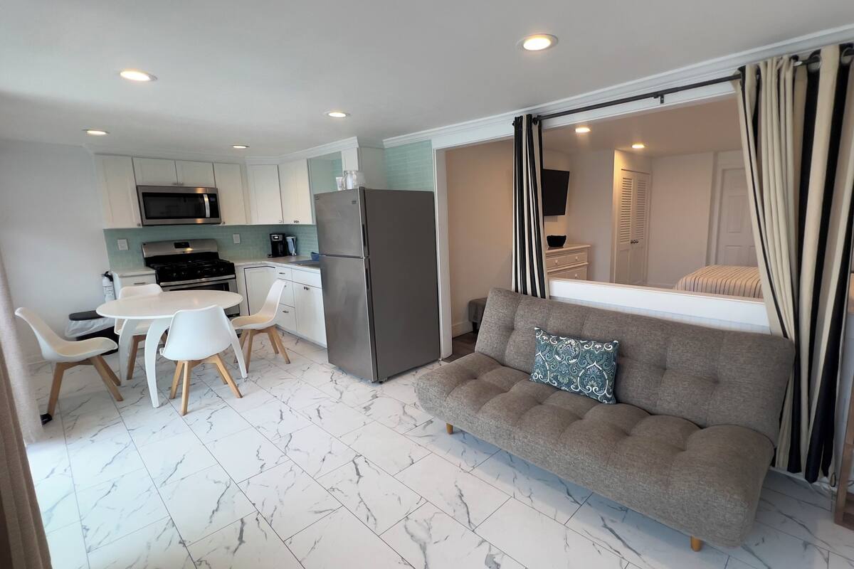 A compact living space with a kitchenette, a sofa with a patterned pillow, and a dining area with white chairs.
