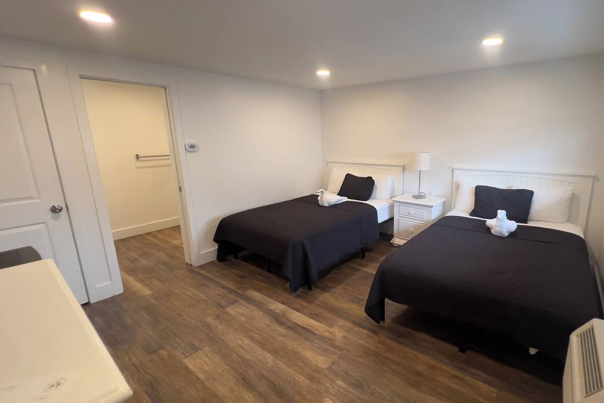 A room with two beds, a bedside table with a lamp, a heater, and a window.