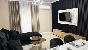 Une chambre d’hôtel moderne avec un canapé d’angle noir, une table ronde dorée, une table à manger dressée pour deux personnes, un téléviseur à écran plat fixé à un mur noir et un climatiseur mural.