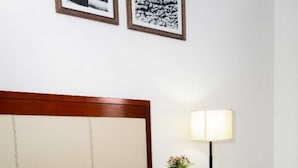 Una cama bien hecha con ropa de cama blanca y gris, un cabecero de madera, una mesita de noche con una lámpara y un pequeño jarrón con flores.