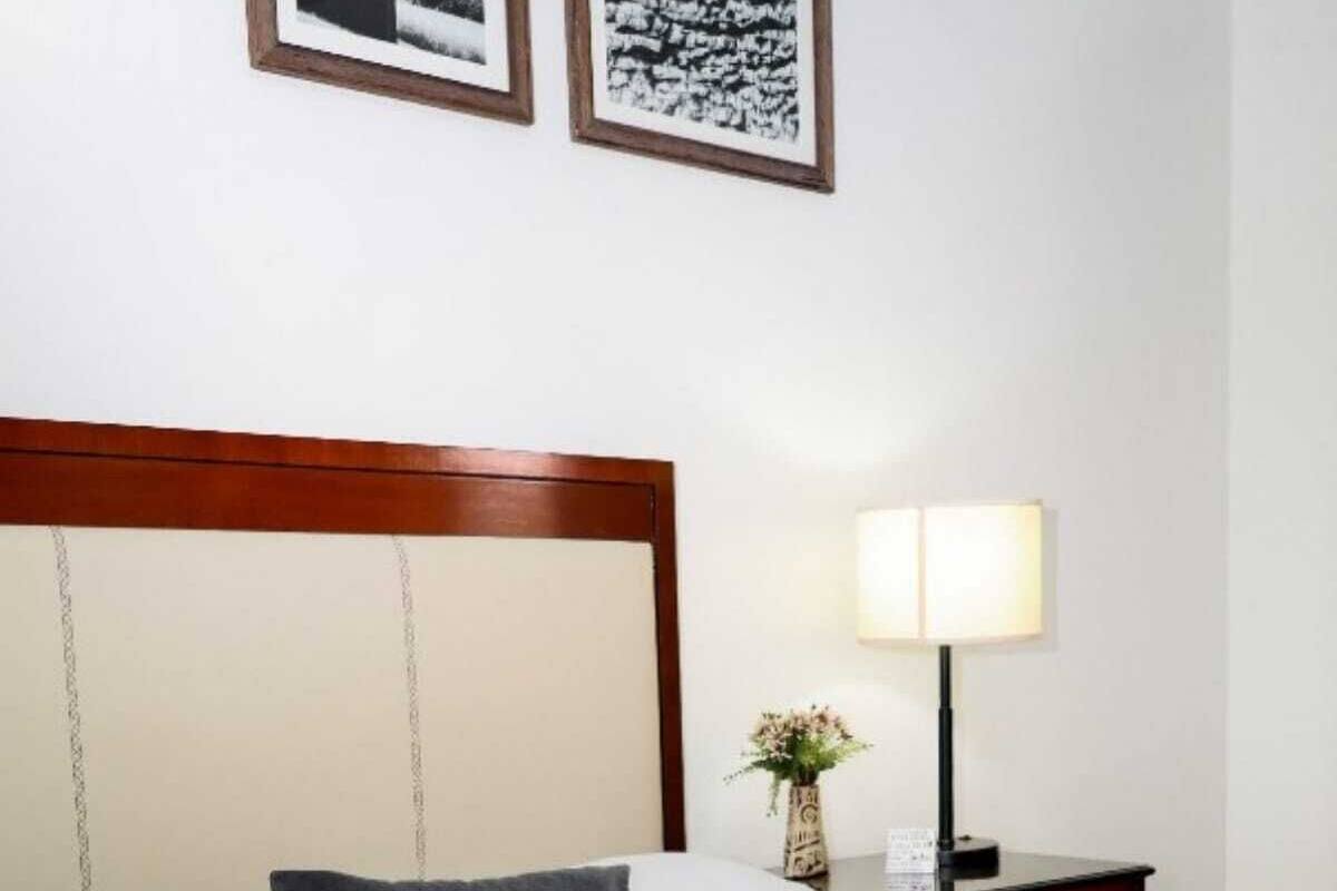 Una cama bien hecha con ropa de cama blanca y gris, un cabecero de madera, una mesita de noche con una lámpara y un pequeño jarrón con flores.