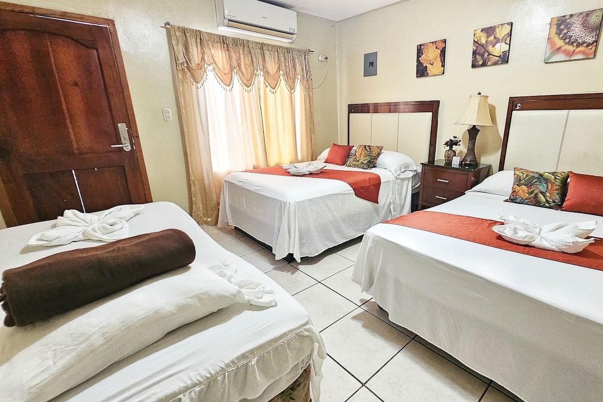 Habitación de hotel con dos camas, una puerta de madera, una ventana con cortinas, una mesita de noche, una lámpara y decoración en la pared.