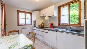 A compact kitchen with white cabinets, a dining table with a floral tablecloth, and a window with a view of greenery. - LEGENT - GRESY SUR AIX (GRESY SUR AIX)