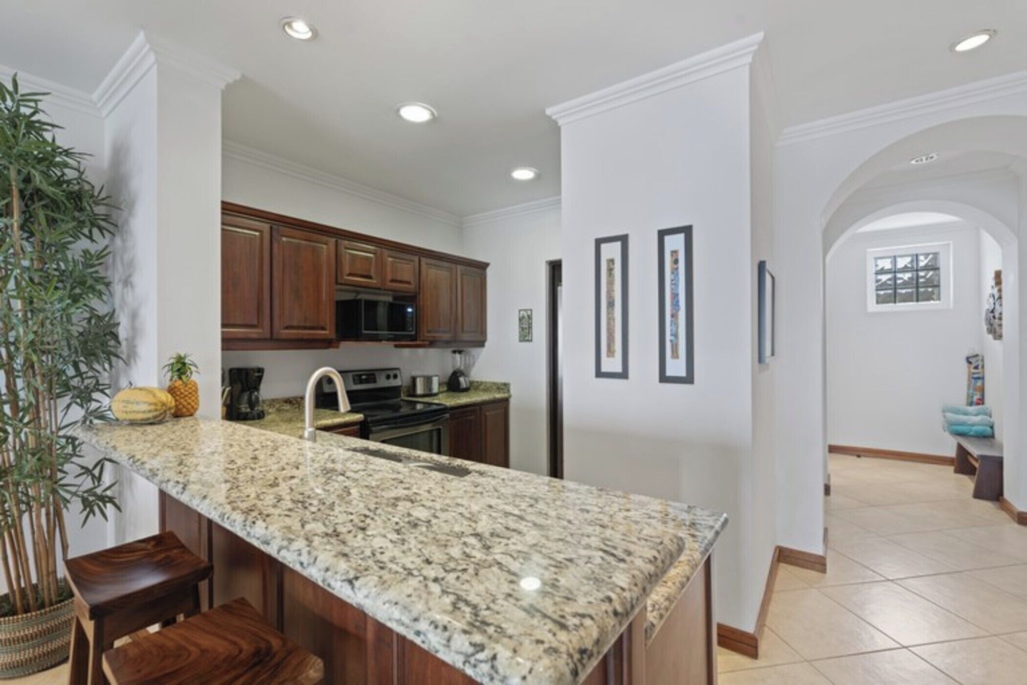 A kitchen with granite countertops, wooden cabinets, a microwave, and a refrigerator. There is a plant and a fruit bowl on the counter.