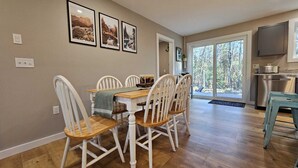Un coin repas avec une table en bois et des chaises blanches, une cuisine dotée d’un réfrigérateur en acier inoxydable et une grande fenêtre donnant sur la forêt.