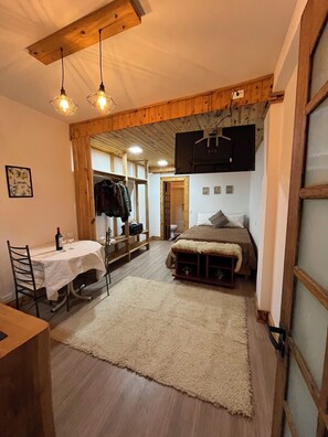 A cozy room with a bed, a table with chairs, a TV mounted on the wall, and a wooden ceiling.
