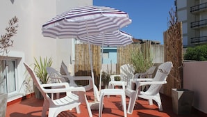 Un patio con sillas de plástico blancas y un paraguas a rayas, rodeado de plantas y una cerca de bambú.